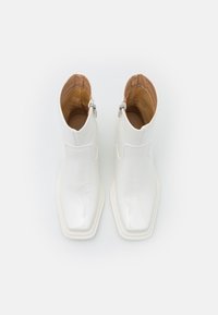 Botas de tobillo de charol blanco con forma de punta, con cremallera lateral y un forro interior en contraste de color marrón claro. Suela plana blanca.