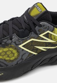 Sort og grå sneaker med gule accenter, der har en struktureret mesh-overdel, snørebåndslukning og et iøjnefaldende logo-design på siden.