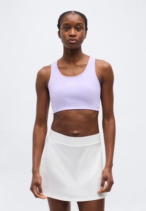 Αθλητική γυναίκα που φοράει ανοιχτό μωβ κοντό τοπ και λευκή φούστα, στέκεται μπροστά σε απλό ανοιχτό γκρι φόντο.