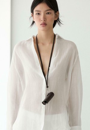 Młoda kobieta ubrana w przezroczystą białą koszulę zapinaną na guziki, z czarną smyczą trzymającą małe cylindryczne czarne etui na szyi.