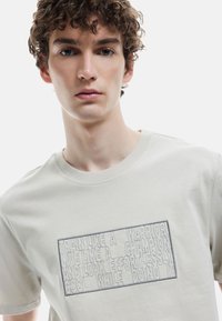 Lys grå bomulds t-shirt med et rektangulært broderet tekstdesign i mørkere grå. Klassisk rund hals med korte ærmer.