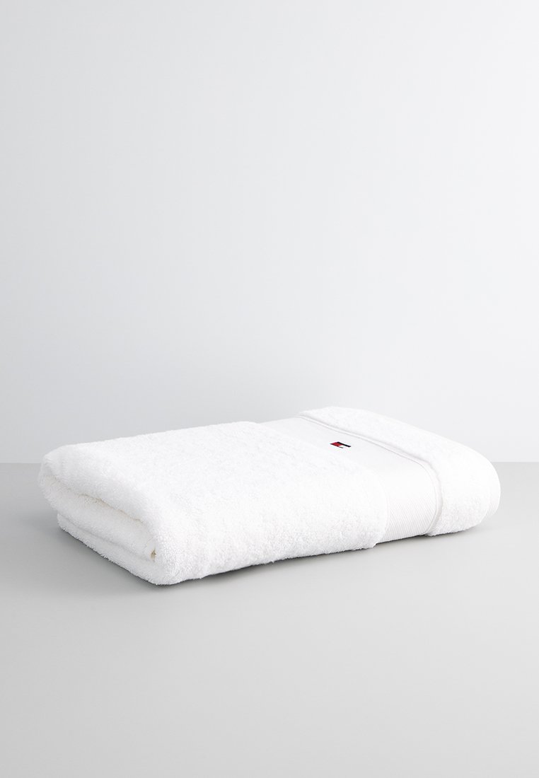 Witte katoenen handdoek met een zachte, pluche textuur, voorzien van een gestikt labelaccent in rood en marineblauw. Netjes gevouwen op een lichtgrijze ondergrond.