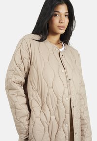 Giacca trapuntata beige con colletto rotondo, bottoni a pressione e motivo ondulato testurizzato. Realizzata in materiale morbido e leggero.