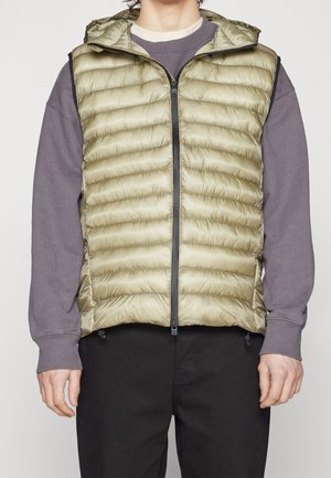 Mężczyzna ubrany w jasnooliwkową pikowaną kamizelkę bez rękawów na szarym długim rękawie i czarnych spodniach, stojący na jednolitym tle.