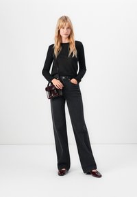 Γυναίκα με ξανθά μαλλιά, ντυμένη με μαύρο μπλουζάκι με μακριά μανίκια, μαύρα παντελόνια wide-leg, μπορντό παπούτσια και μια μικρή μπορντό τσάντα crossbody, να στέκεται μπροστά σε άσπρο φόντο.