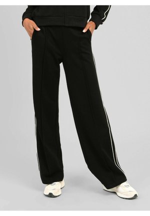 Personne portant un pantalon noir à jambes larges avec des bandes blanches sur les côtés et des baskets blanches, debout les mains dans les poches devant un fond uni.