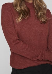 Femme aux cheveux blonds ondulés portant un pull en maille couleur rouille à manches longues et un pantalon noir, les bras croisés à la taille.