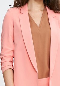 Jasnoróżowy żakiet o strukturze, w połączeniu z brązowym topem z dekoltem w serek. Żakiet ma podwinięte rękawy i dopasowany krój.