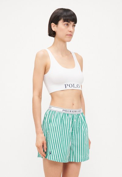 Balta kokvilnas sporta krūšturis ar tumši zilā "POLO" logotipu, apvienots ar zaļiem un baltiem svītrainiem šortiem, kuriem ir elastīga jostasvieta un logotipa detaļa.