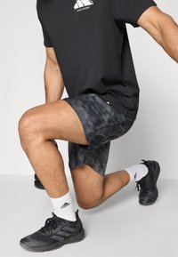 T-shirt de sport noir avec logo blanc, shorts à motifs noir et gris, chaussettes blanches et chaussures de sport noires avec des détails texturés.