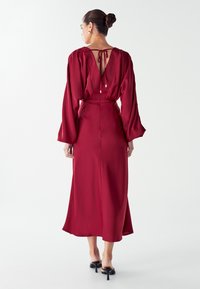 Moteris su susuktomis plaukais, vilkinti ilgomis rankovėmis, raudona midi suknelė su atviru nugaros kirpimu ir juodais aukštakulniais sandalais, stovi ant balto fono.