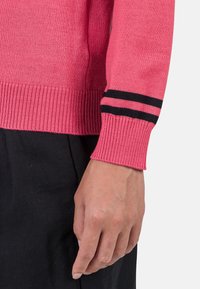 Różowy sweter z dzianiny z prążkowanymi mankietami, ozdobiony czarnymi paskami, ukazujący teksturę materiału oraz zbliżenie na dłoń opierającą się na czarnej odzieży.