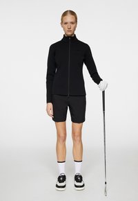 Svart zip-up tröja, svarta shorts, vita strumpor och svarta skor. Golfklubba hålls i ena handen, med en vit handske på den handen.