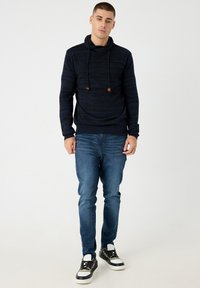 Sudadera con capucha de rayas marineras, con cuello vuelto y cordones, combinada con vaqueros azules y zapatillas negras con detalles en blanco.