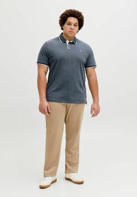 Άνδρας με σγουρά μαλλιά που φοράει σκούρο γκρι πόλο μπλουζάκι, μπεζ παντελόνι και λευκά αθλητικά παπούτσια, στέκεται μπροστά σε ένα απλό φωτεινό φόντο.