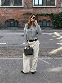 Sivý sveter na gombíky, krémové nohavice so širokými nohavicami, čierny opasok a malá čierna kabelka. Čierne slnečné okuliare. Mestské prostredie s tehlovou stenou v pozadí.