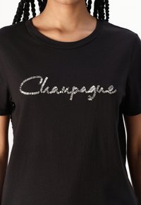 Černé bavlněné tričko s texturou stříbrného nápisu "Champagne" na hrudi, klasickým kulatým výstřihem a krátkými rukávy.