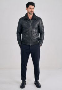 Man staat met handen in de zakken, draagt een zwarte leren jas, donkere broek en zwarte laarzen tegen een effen lichte achtergrond.