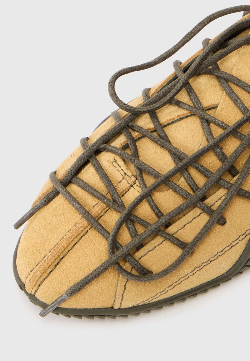 Chaussure en daim beige avec des lacets noirs entrecroisés sur fond blanc, montrant des coutures détaillées et le bord de semelle en caoutchouc.