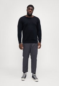 Pulover negru tricotat, pantaloni cargo gri și adidași negri cu glezna înaltă. Puloverul are un decolteu rotund și mâneci ajustate.
