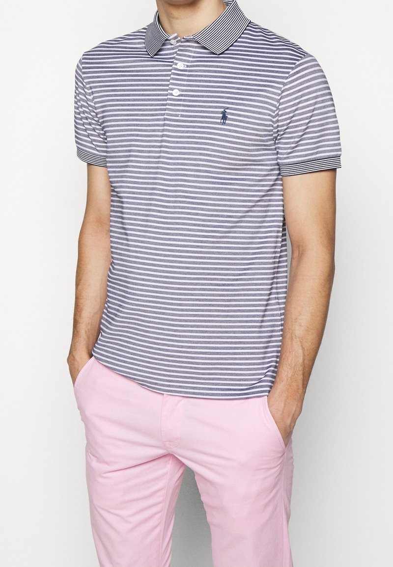 Polo rayé en bleu marine et blanc, avec un col bleu marine uni. Porté avec un pantalon rose clair, affichant des manches courtes et une coupe classique.