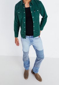 Camicia di velluto a coste verde, canottiera nera, jeans patchwork azzurri e stivali marroni alla caviglia. Presenta tasche con bottoni e tessuto strutturato.