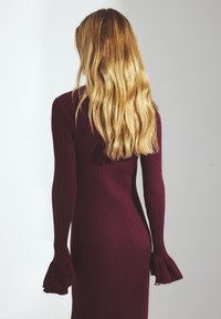 Abito a maniche lunghe, in borgogna, a coste, con polsini svasati, silhouette aderente e tessuto testurizzato. La vista posteriore mostra lunghi capelli mossi.