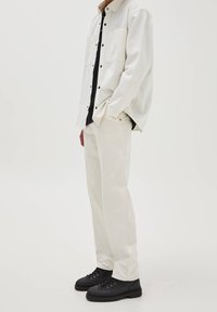 Biała koszula zapinana na guziki z dwoma kieszeniami na piersi, noszona na czarnej koszuli, w połączeniu z off-white spodniami i czarnymi butami na sznurowadła. Gładka tekstura materiału.