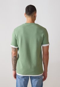 T-shirt vert à manches courtes avec un motif texturé, col et poignets côtelés blancs, vue de dos, porté avec un jean bleu.