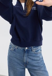 Sudadera azul marino con cremallera, cuello alto y puños acanalados, combinada con jeans de tiro alto azul claro con cierre de botones.