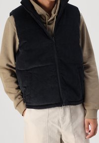 Chaleco de pana negro con cierre de cremallera, que presenta un cuello alto y bolsillos laterales, superpuesto a una sudadera beige con capucha.