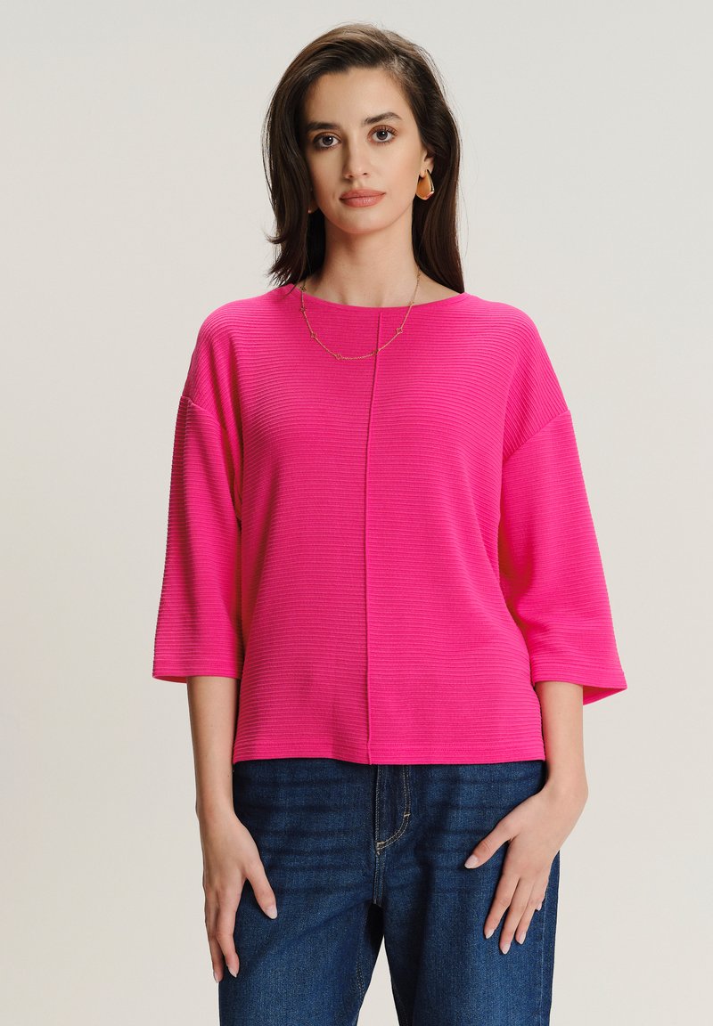 Kobieta w jasnoróżowym, prążkowanym topie z trzema ćwiartkami rękawów i niebieskich dżinsach, stojąca na tle jednolitego jasnego tła.