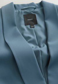 Giacca in raso blu con colletto a scialle. Presenta una trama liscia. Etichetta con il nome del marchio "NEXT" e dettagli per la cura.