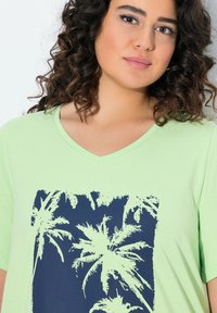 T-shirt verde chiaro con scollatura a V caratterizzata da una stampa grafica di palma blu scuro. Maniche corte e texture del tessuto morbida.