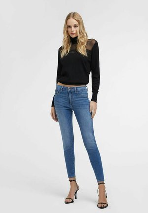 Donna che indossa una maglia nera a maniche lunghe con scollo trasparente, jeans skinny blu e sandali neri con tacco alto, in piedi contro uno sfondo semplice.