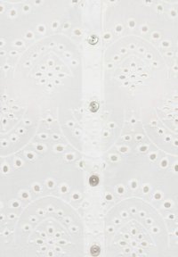 Tissu blanc avec des motifs floraux en broderie ajourée et une rangée verticale de petits boutons ronds aux reflets nacrés.