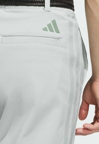 Light gray athletic pants with a smooth texture, side stripes, and a rear pocket featuring a green Adidas logo.