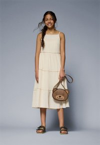 Cream-colored, tiered dress with spaghetti straps, paired with a brown bear-shaped handbag and green sandals with black buckles.