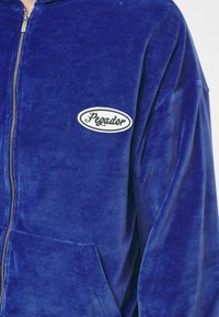 Sudadera de terciopelo azul con cremallera, bolsillo frontal y un parche ovalado blanco que muestra "Pegador" en escritura negra. Textura suave y corte relajado.
