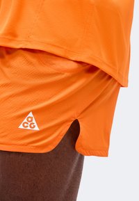 Κοντινή λήψη ενός ατόμου που φοράει φωτεινά πορτοκαλί αθλητικά σορτς και αντίστοιχο μπλουζάκι με λευκό λογότυπο ACG στα σορτς.