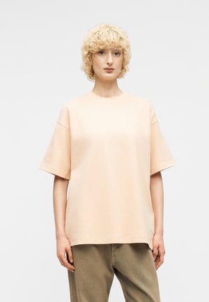 Άτομο με κοντά, σγουρά ξανθά μαλλιά που φοράει ένα χαλαρό, παλ ροδακινί κοντομάνικο πουκάμισο και παντελόνι σε ελιά πράσινο χρώμα, στέκεται μπροστά σε απλό φόντο.