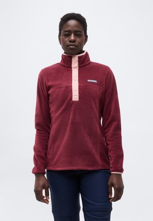 Persoon die een kastanjebruine fleece trui draagt met roze drukknoopjes en een donkerblauwe broek, staand tegen een effen lichte achtergrond.