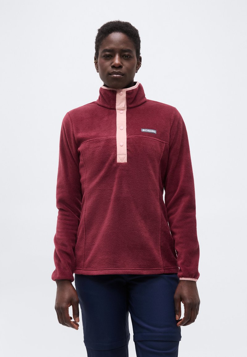 Persoon die een kastanjebruine fleece trui draagt met roze drukknoopjes en een donkerblauwe broek, staand tegen een effen lichte achtergrond.