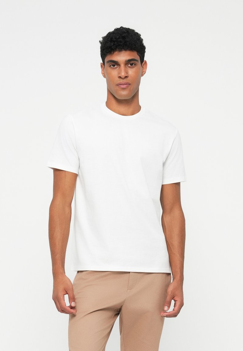 Junger Mann mit lockigem Haar, der ein einfaches weißes kurzärmliges T-Shirt und beige Hosen trägt, steht vor weißem Hintergrund.