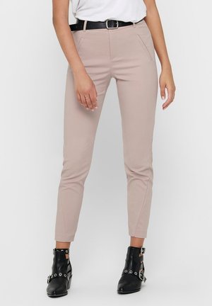 Frau, die beige, eng anliegende Hose mit vorderen Taschen, schwarzen Gürtel, weißen Top und schwarze Stiefeletten mit silbernen Schnallen und Ösen trägt.
