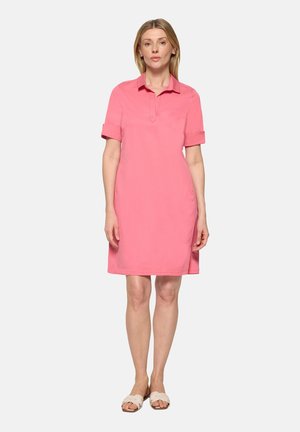 Erwachsene Frau steht, trägt ein knielanges rosa Kleid mit kurzen Ärmeln und Kragen sowie weiße Sandalen mit offenem Zehenbereich, neutraler Hintergrund.