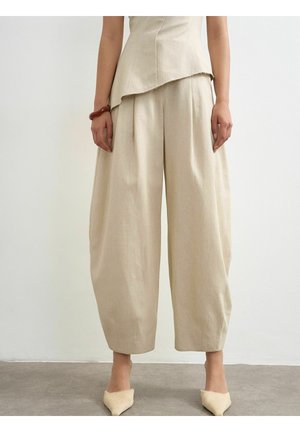 Vrouw draagt losse beige cropped broek, bijpassende top, bruine armband en beige puntige platte schoenen, staand op een grijze vloer.