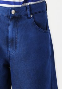Blå denim bukser har en klassisk passform med en frontlomme, metalknaplukning og synlig stikning. Glat tekstur.