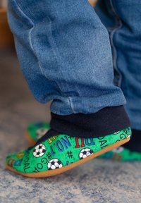 Groene stoffen slippers met patroon van voetballen en kleurrijke tekst. Zacht materiaal met een flexibele zool, gedragen met blauwe spijkerbroeken.