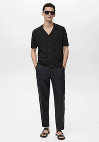 Black short-sleeve button-up shirt with ribbed texture, paired with dark trousers and black sandals, worn with sunglasses.